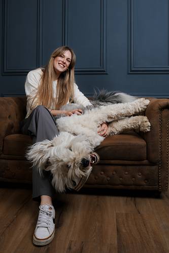 Young woman smiling while cuddling her fluffy Old English Sheepdog on a brown vintage sofa. The dog lies upside down playfully during a cozy pet photoshoot in the studio.